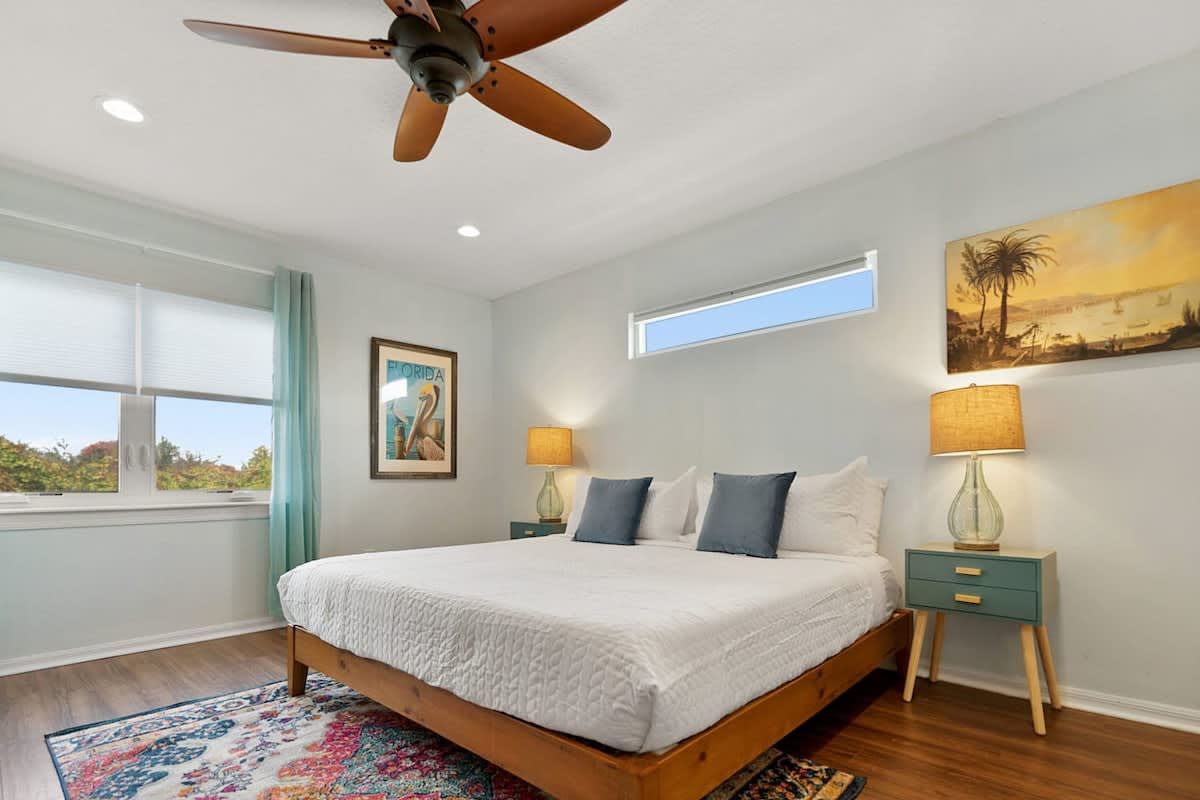 A bedroom with a bed, two bedside tables, a ceiling fan, a window, and a painting on the wall.