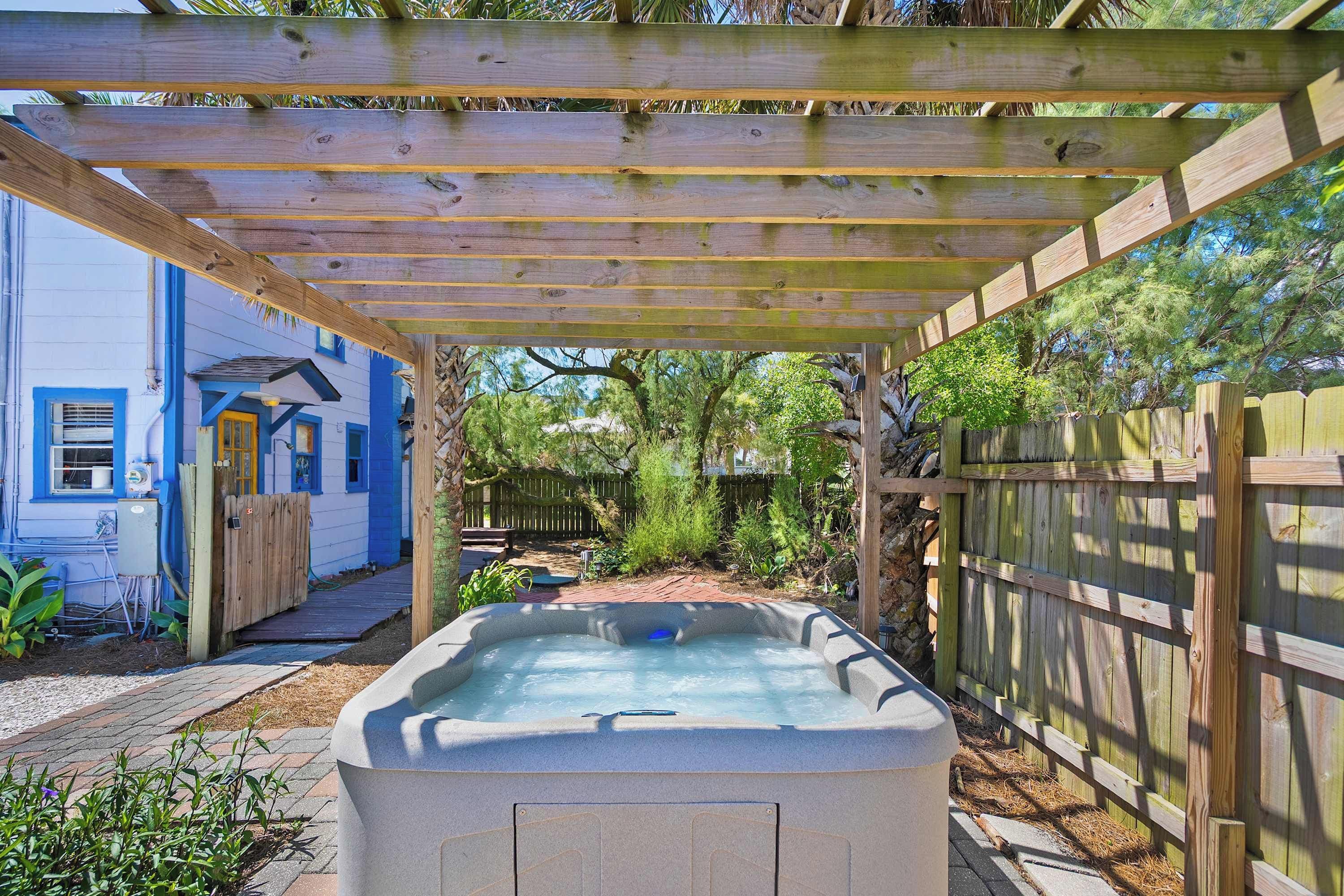 Un bain à remous sous une pergola en bois, dans un jardin avec une maison bleue et une clôture en bois.