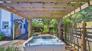 Un bain à remous sous une pergola en bois, dans un jardin avec une maison bleue et une clôture en bois.