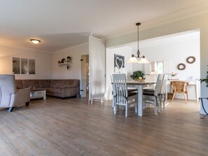 A spacious living area with a sofa, dining table, and chairs, a kitchen in the background, and a ceiling light fixture. - Apartment 90 in the Stella Guesthouse (St. Peter-Ording)