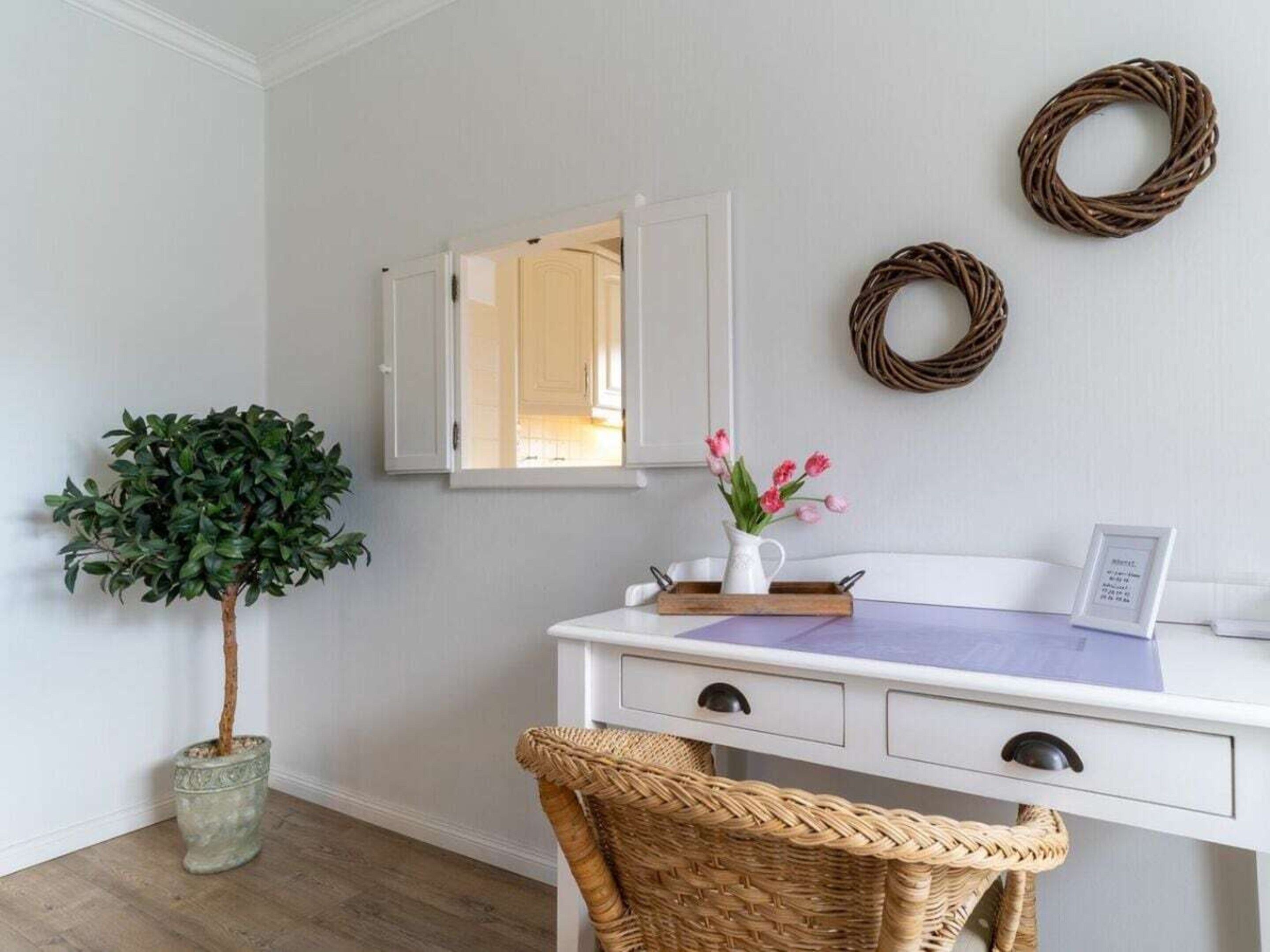 A room with a white desk, a potted plant, a wicker chair, a mirror, and two wall-mounted wreaths.