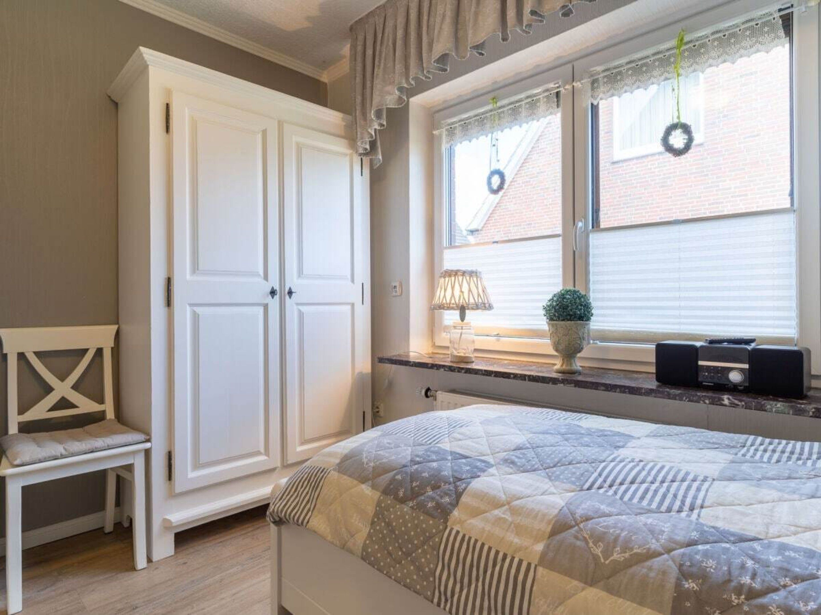 Un dormitorio con cama, armario blanco, ventana con cortinas, mesita de noche con lámpara y planta en maceta.