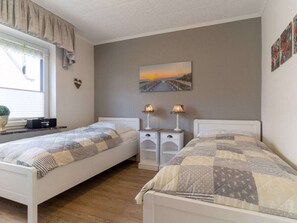 A bedroom with two single beds, a bedside table with lamps, a window with curtains, a wall-mounted picture, and a small plant on the windowsill. - Apartment 90 in the Stella Guesthouse (St. Peter-Ording)