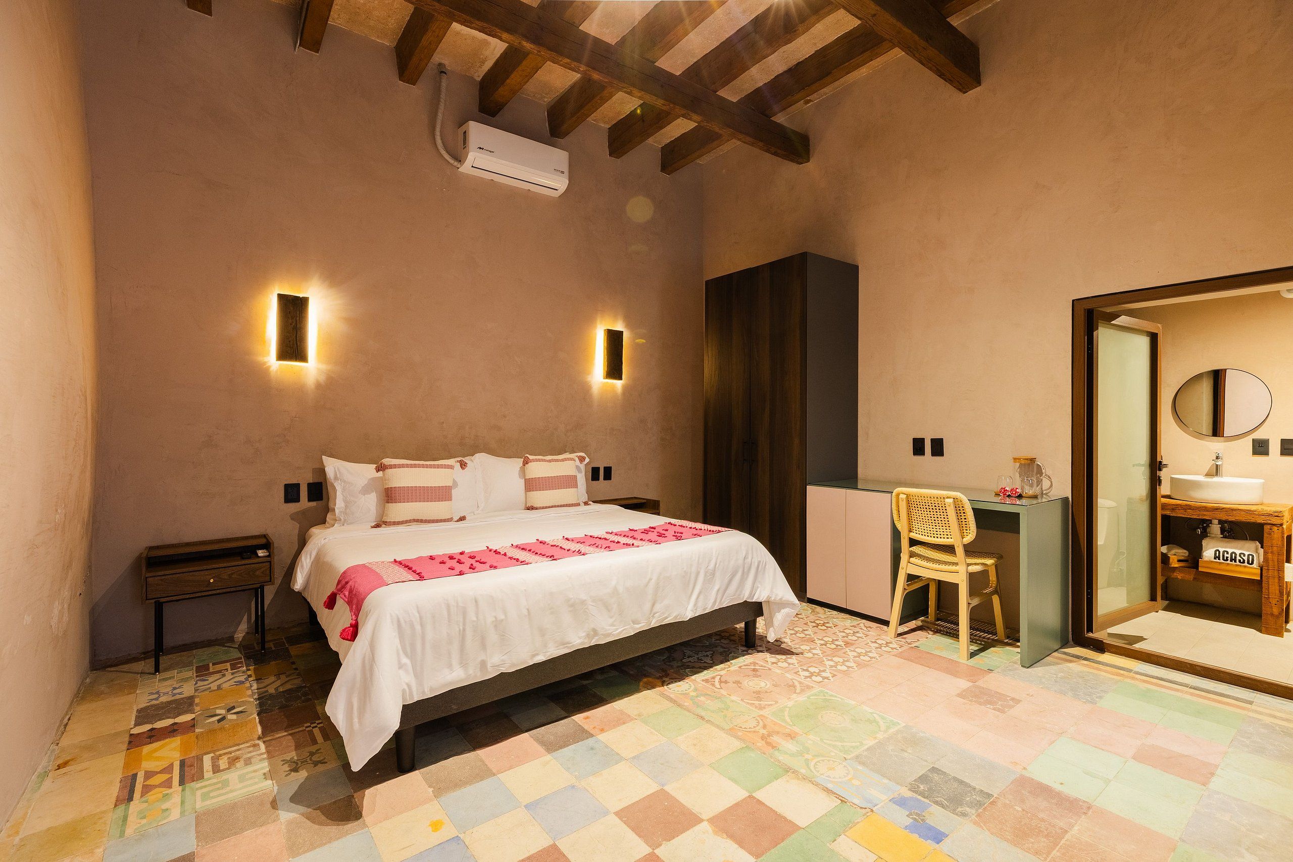 A bedroom with a bed, bedside tables, a chair, a mirror, and a bathroom with a sink and mirror.