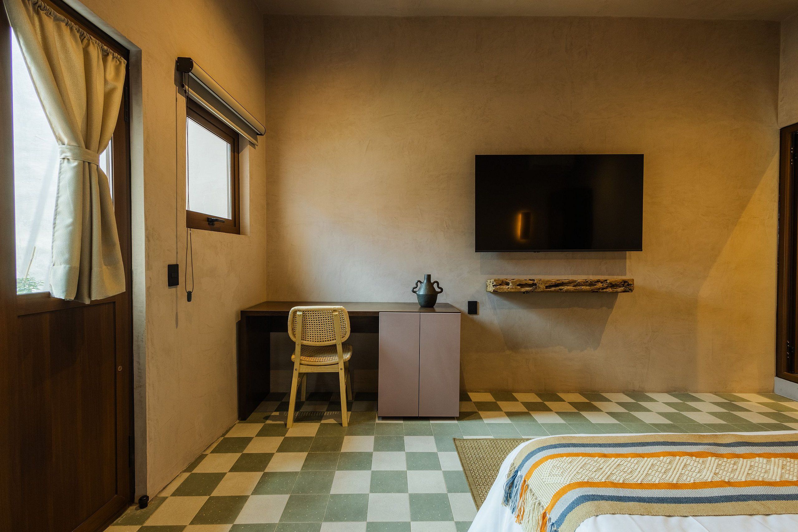 A room with a checkered floor, a bed, a desk with a chair, a television mounted on the wall, and a window with curtains.