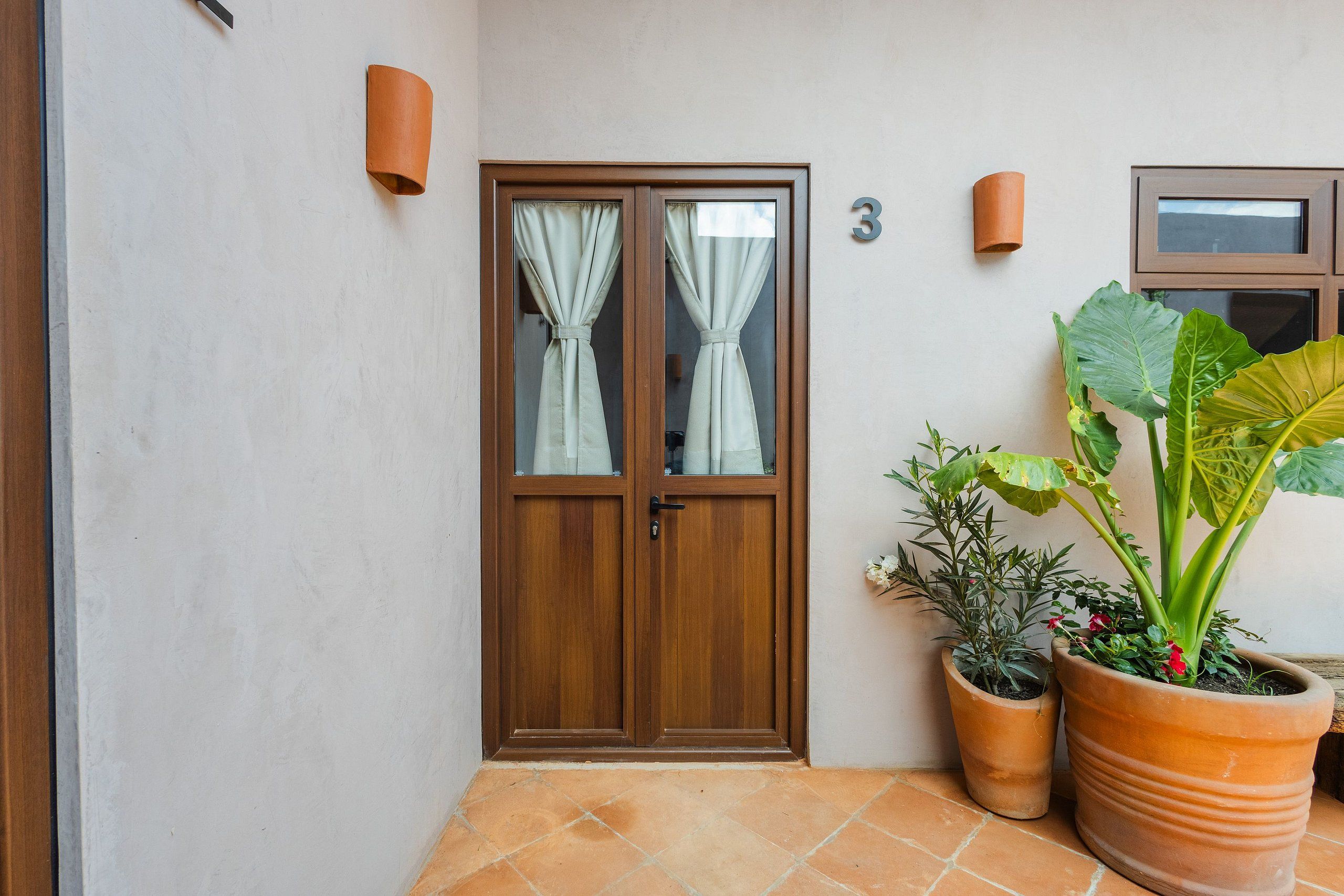 A wooden door with glass panels and curtains, a number '3' sign, and potted plants.