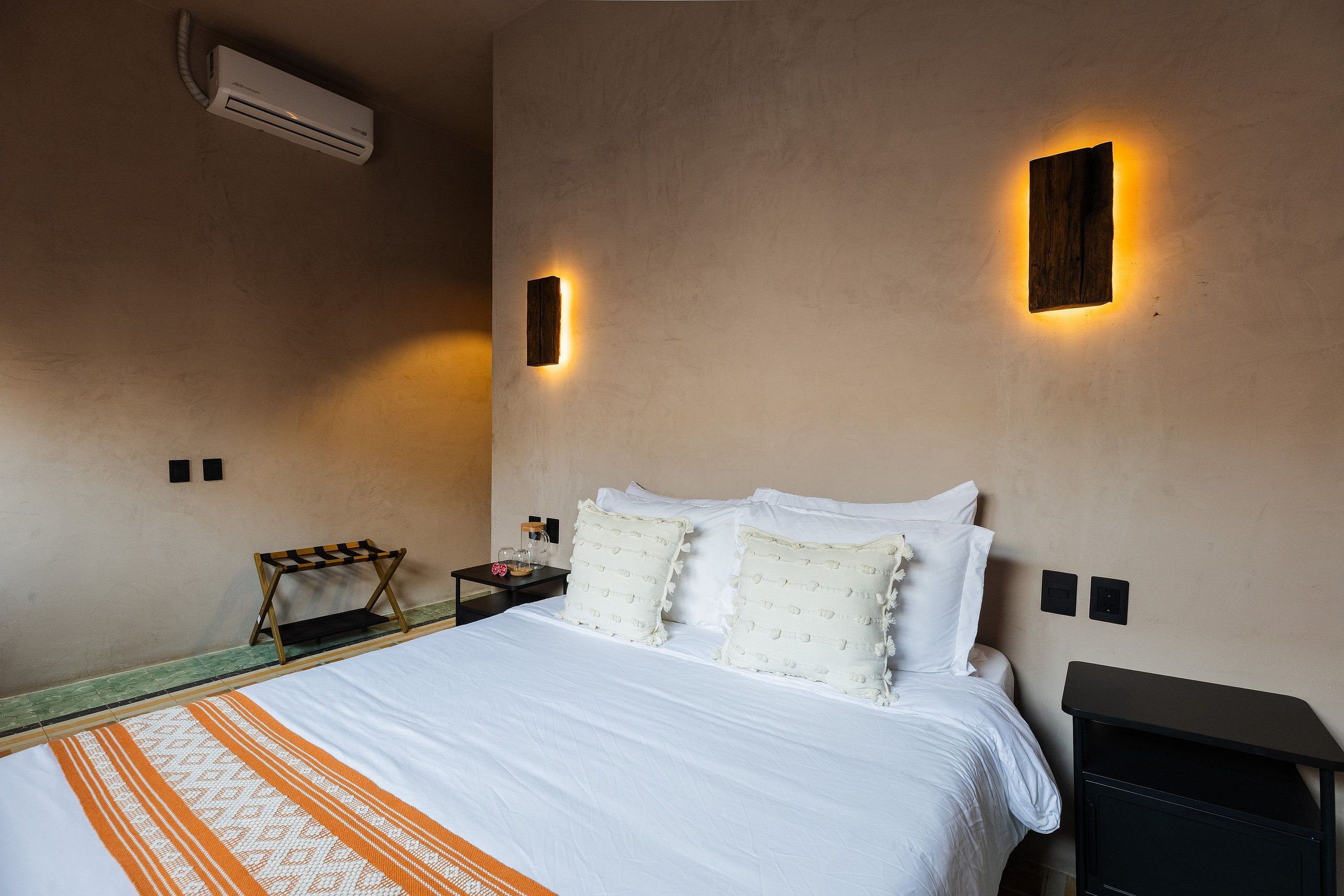 A hotel room with a bed, bedside tables, an air conditioning unit, and wall-mounted lighting.