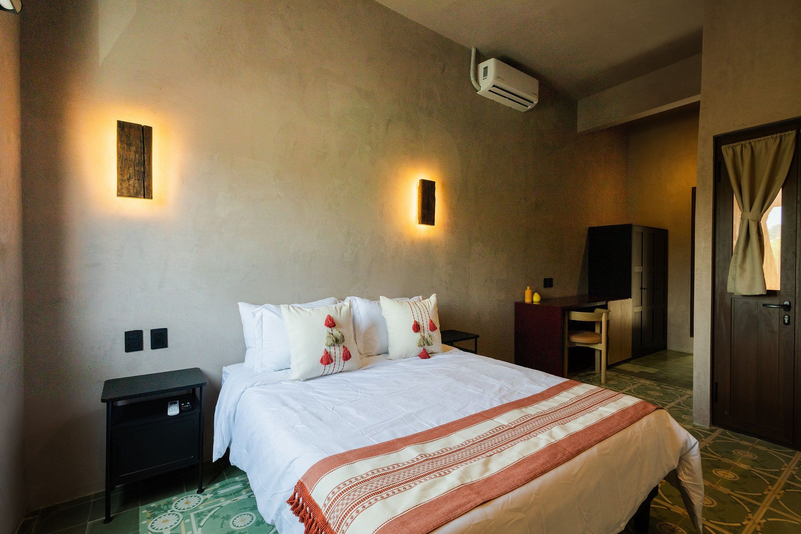 A hotel room with a bed, bedside tables, a desk, an air conditioning unit, and a door with curtains.