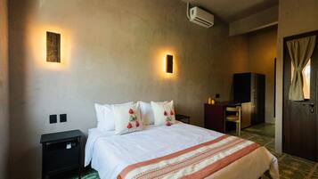 A hotel room with a bed, bedside tables, a desk, an air conditioning unit, and a door with curtains.
