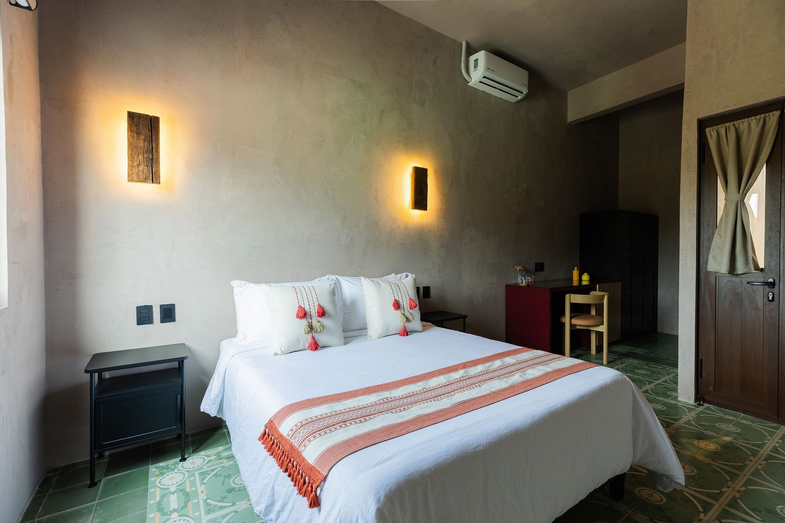 A hotel room with a bed, bedside tables, an air conditioning unit, a wall-mounted light, and a door with curtains.