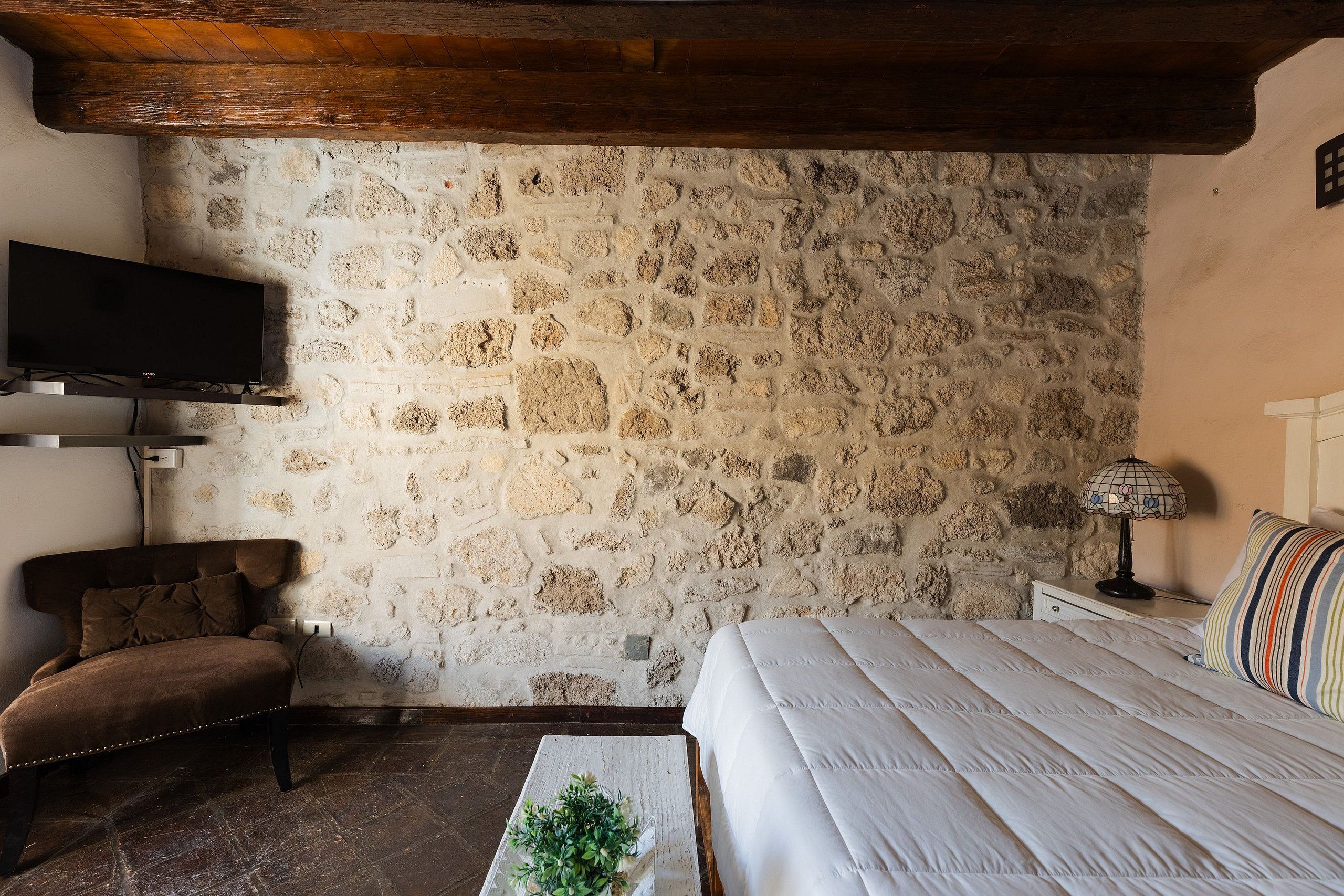 A bedroom with a stone wall, a bed, a TV mounted on the wall, a chair, and a bedside table with a lamp.