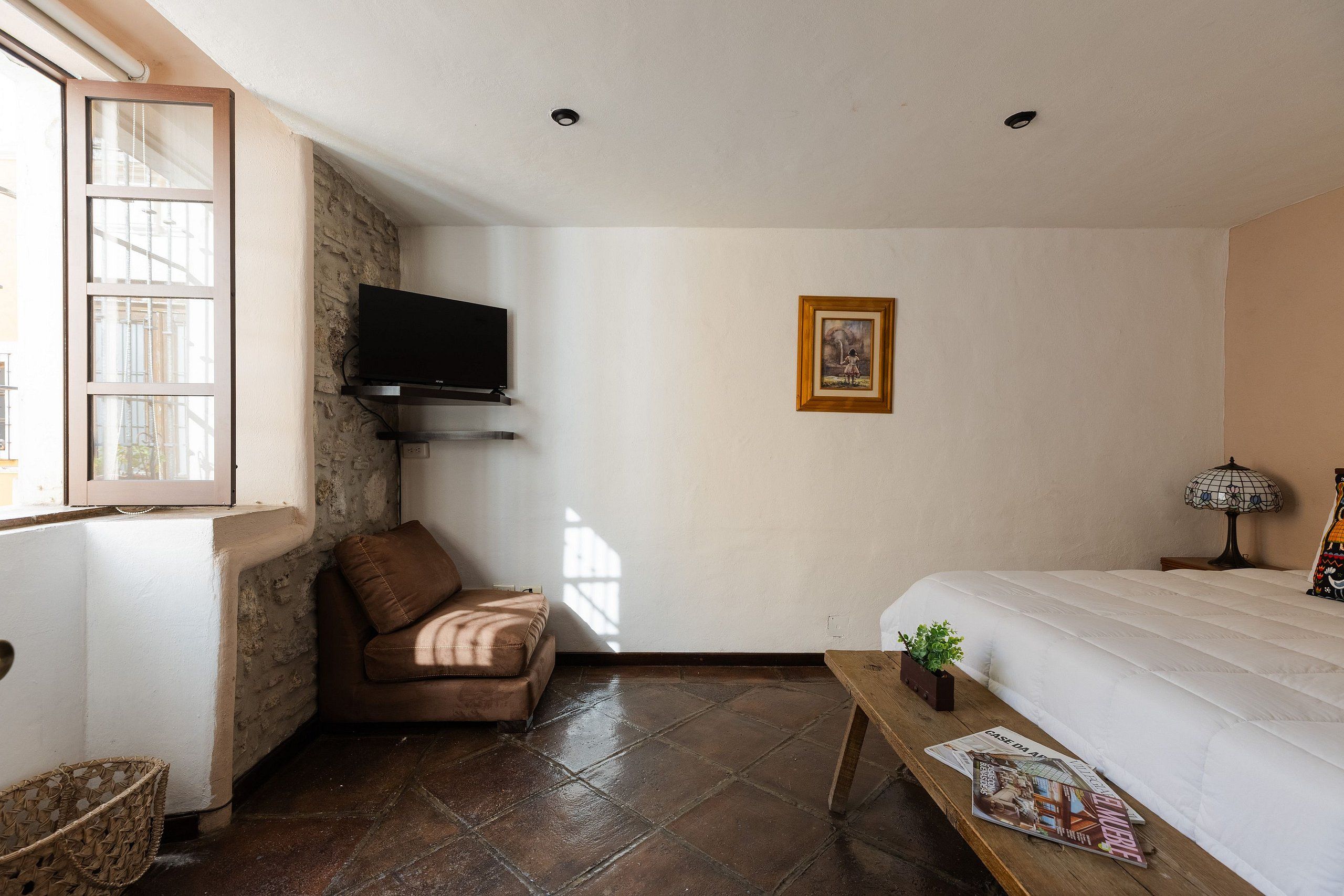 A room with a bed, a television mounted on a brick wall, a framed picture on the wall, a window with blinds, and a wooden table with magazines.