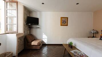 A room with a bed, a television mounted on a brick wall, a framed picture on the wall, a window with blinds, and a wooden table with magazines.