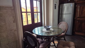 Une cuisine avec une table ronde en verre, des chaises en osier, un réfrigérateur et une vue sur l’extérieur par une fenêtre qui donne sur une porte.