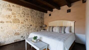 A bedroom with a stone wall, a wooden ceiling, a bed with white bedding, and a small table with a plant.