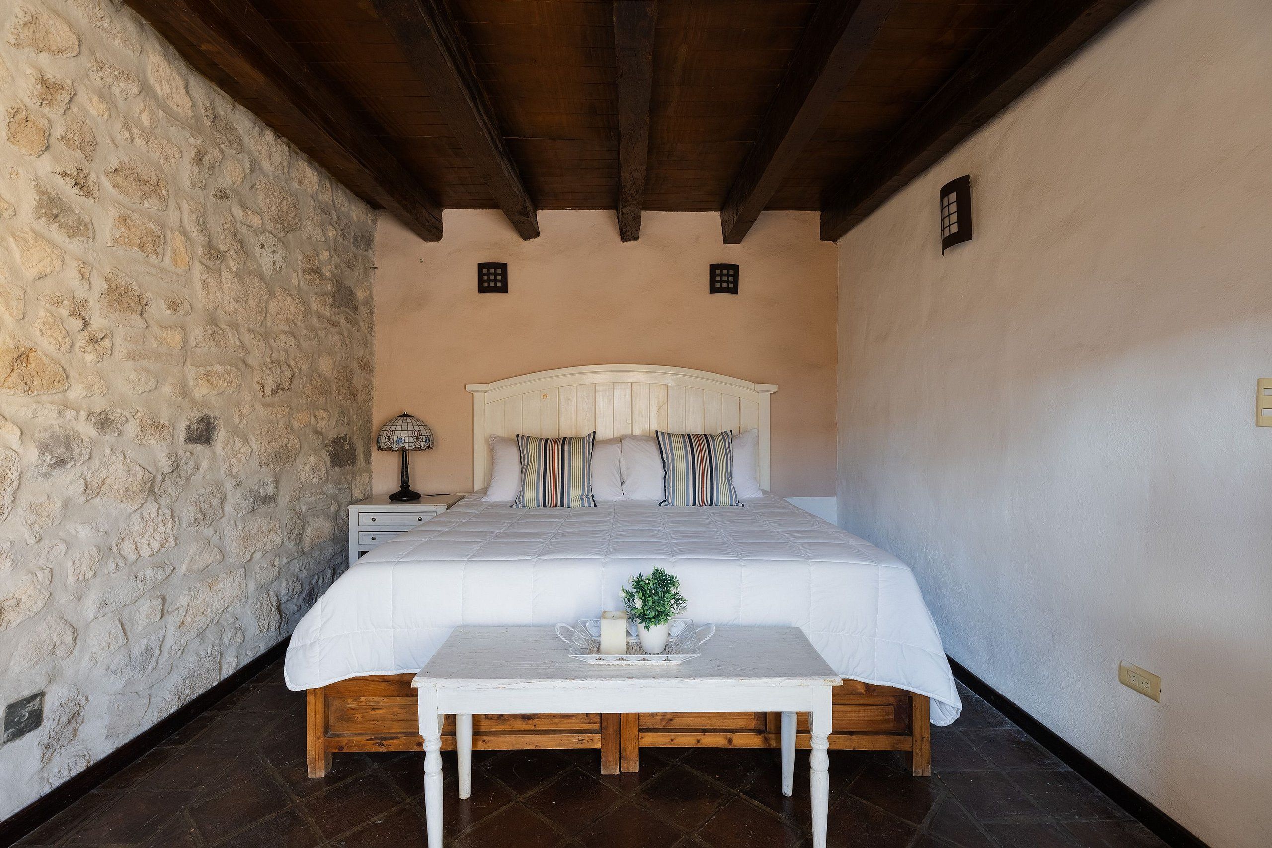 A bedroom with a stone wall, a wooden ceiling, a bed with a white headboard, a bedside table with a lamp, and a small plant on the table.