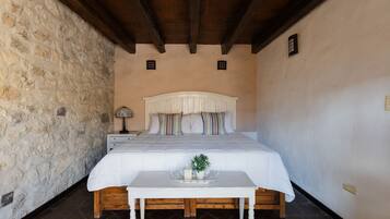A bedroom with a stone wall, a wooden ceiling, a bed with a white headboard, a bedside table with a lamp, and a small plant on the table.