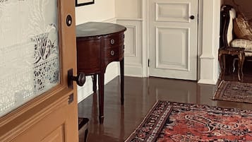 A room with a wooden door, a dark wooden table, a patterned rug, a white door with a double panel, and a hanging light fixture.