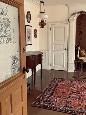 A room with a wooden door, a dark wooden table, a patterned rug, a white door with a double panel, and a hanging light fixture.