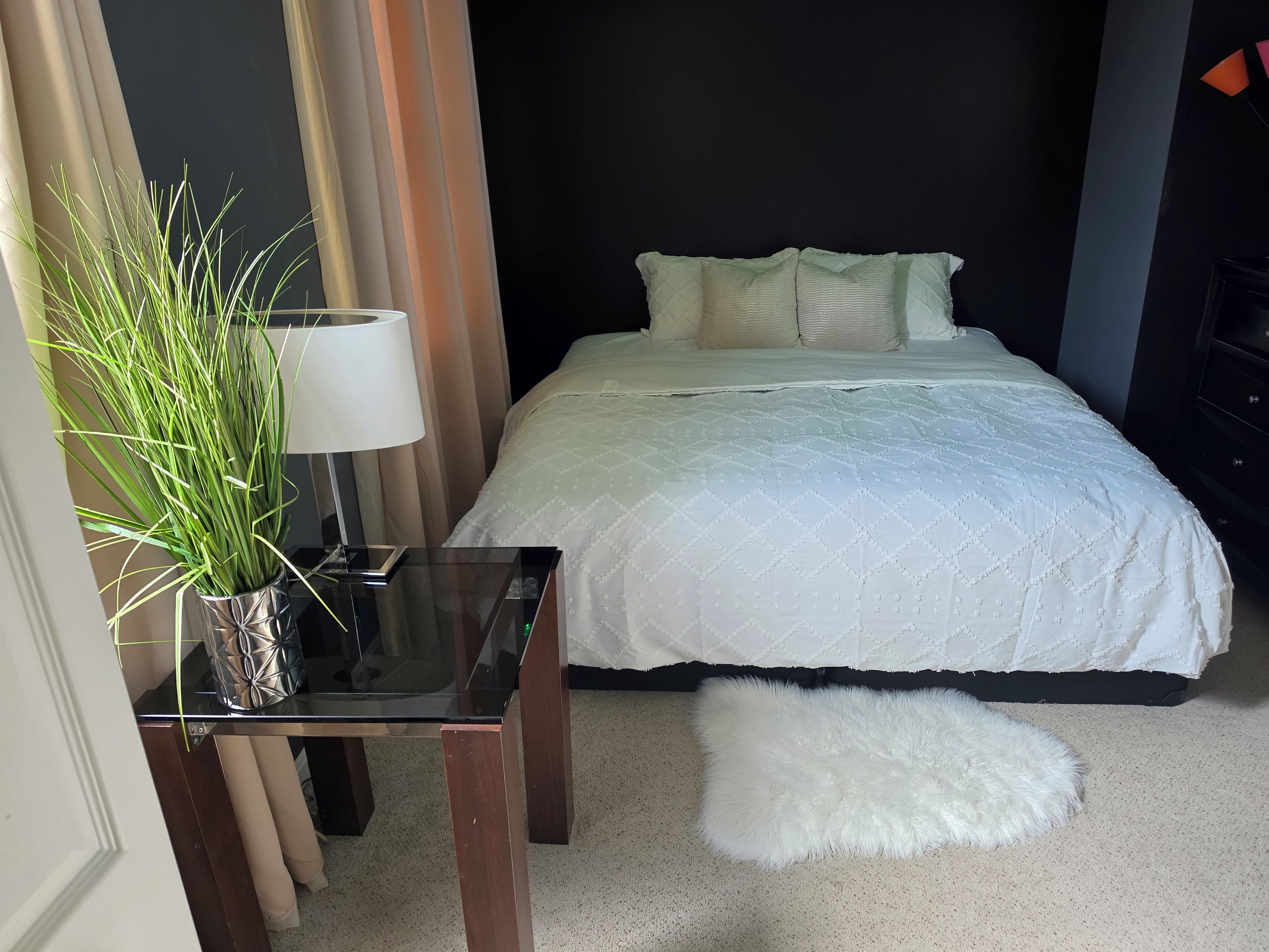 Quarto de hotel com cama, mesa de cabeceira com abajur, planta em vaso e tapete branco felpudo.