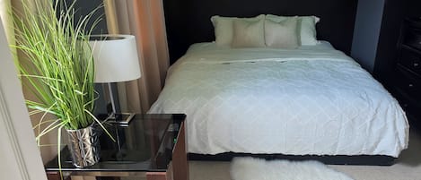 Quarto de hotel com cama, mesa de cabeceira com abajur, planta em vaso e tapete branco felpudo.