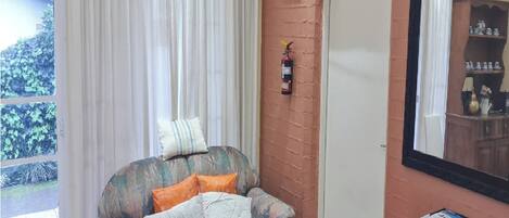 A room with a window, a fire extinguisher mounted on the wall, a chair with cushions, and a table with a stack of magazines.