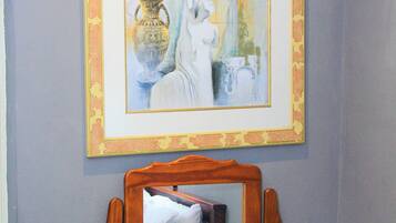 Une œuvre d’art encadrée est accrochée au-dessus d’une commode en bois, sur laquelle se trouvent un miroir, une télécommande et un coussin.
