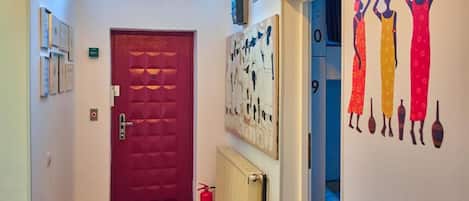 Un pasillo con una puerta roja, un extintor, un radiador y cuadros con figuras vestidas con atuendo tradicional.