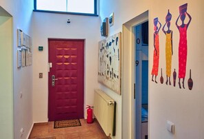 A hallway with a red door, a fire extinguisher, a radiator, and wall art depicting figures in traditional attire.