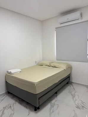 Ein Einzelbett mit Matratze, Kissen und einem zusammengerollten Handtuch in einem Zimmer mit weißen Wänden und Fliesenboden.