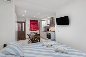 Un dormitorio con cama, una mesa de comedor con sillas, una zona de cocina con armarios y estufa, y un televisor montado en la pared.