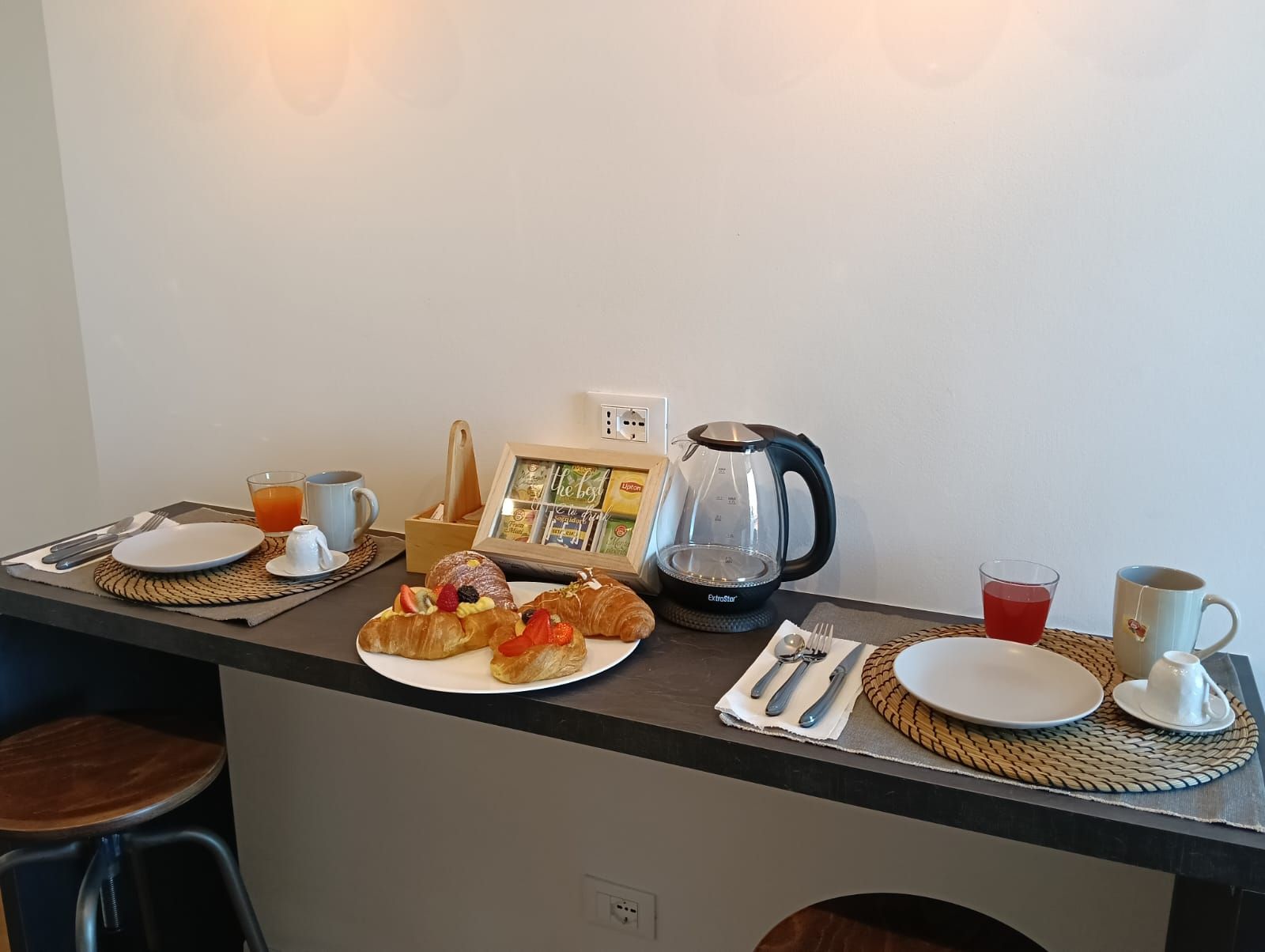 Ein Frühstücksbuffet mit Croissants, Obst, einer Kaffeemaschine und einem gerahmten Bild auf einer Arbeitsplatte.