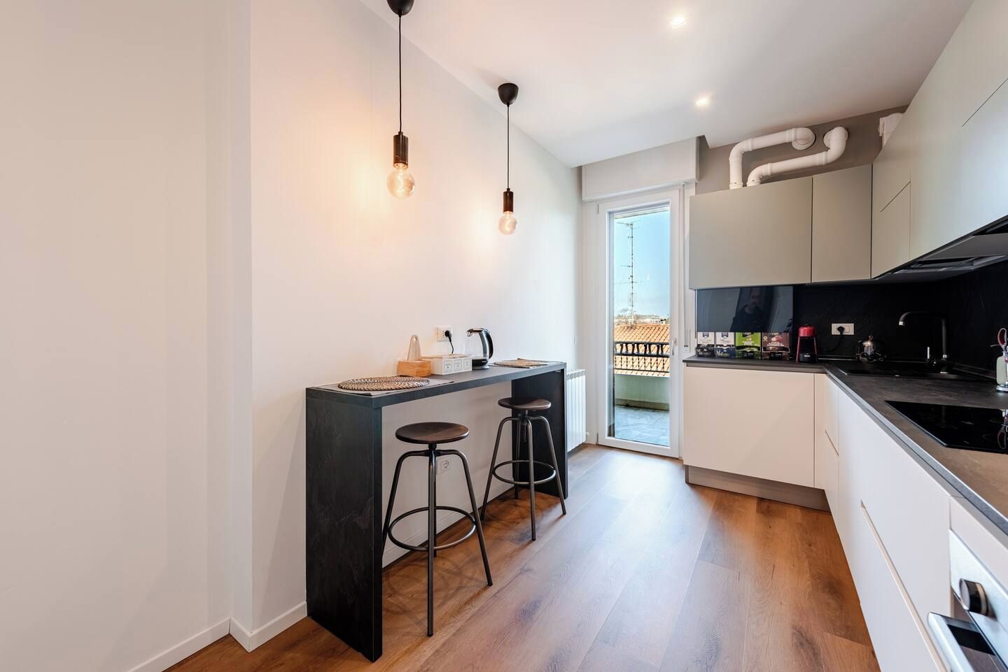 Une cuisine moderne avec un plan de travail noir, deux tabourets de bar et des luminaires suspendus.