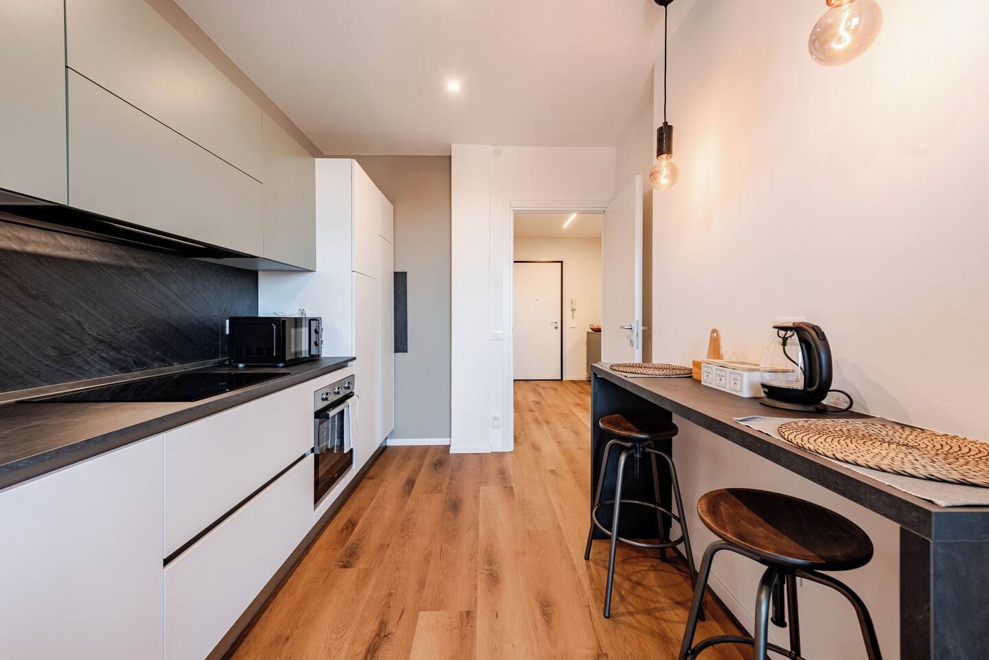 Eine moderne Küche mit schwarzer Arbeitsplatte, Edelstahlgeräten und Holzboden. Auf der Arbeitsplatte befinden sich zwei Hocker und eine Kaffeemaschine.