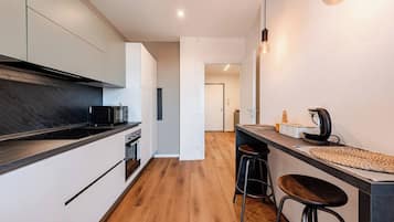 Eine moderne Küche mit schwarzer Arbeitsplatte, Edelstahlgeräten und Holzboden. Auf der Arbeitsplatte befinden sich zwei Hocker und eine Kaffeemaschine.