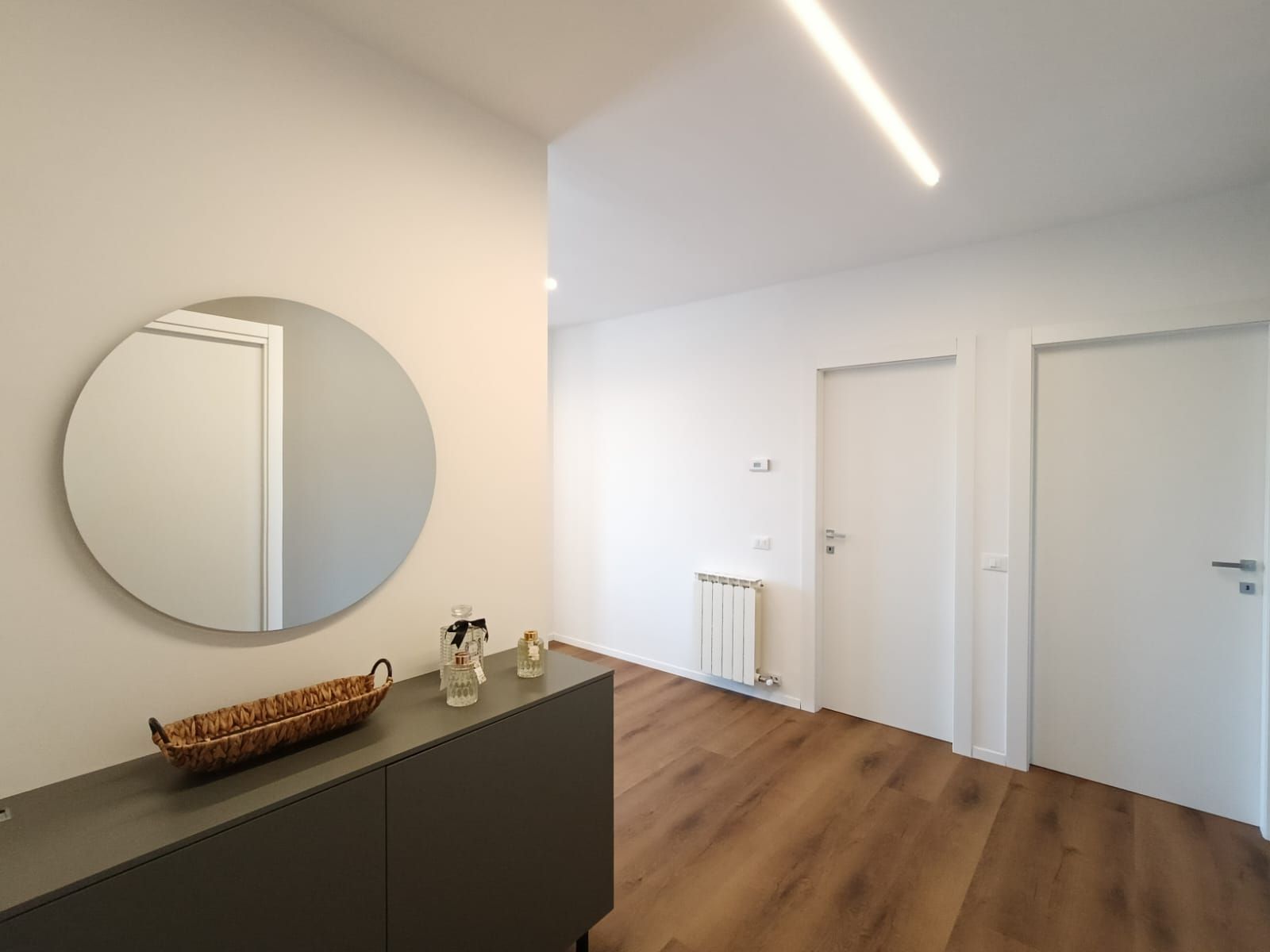 Une chambre moderne avec un grand miroir rond, une commode de couleur sombre et un plancher en bois. On y trouve deux portes blanches et un radiateur au mur.