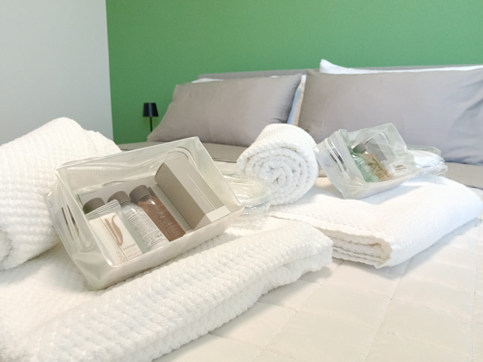 Un letto rifatto con lenzuola bianche, un asciugamano arrotolato e un contenitore di plastica trasparente con articoli da toilette.