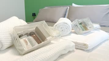 Un letto rifatto con lenzuola bianche, un asciugamano arrotolato e un contenitore di plastica trasparente con articoli da toilette.
