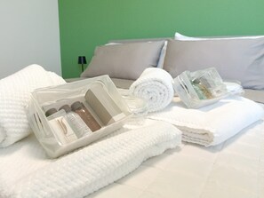 Un letto rifatto con lenzuola bianche, un asciugamano arrotolato e un contenitore di plastica trasparente con articoli da toilette.