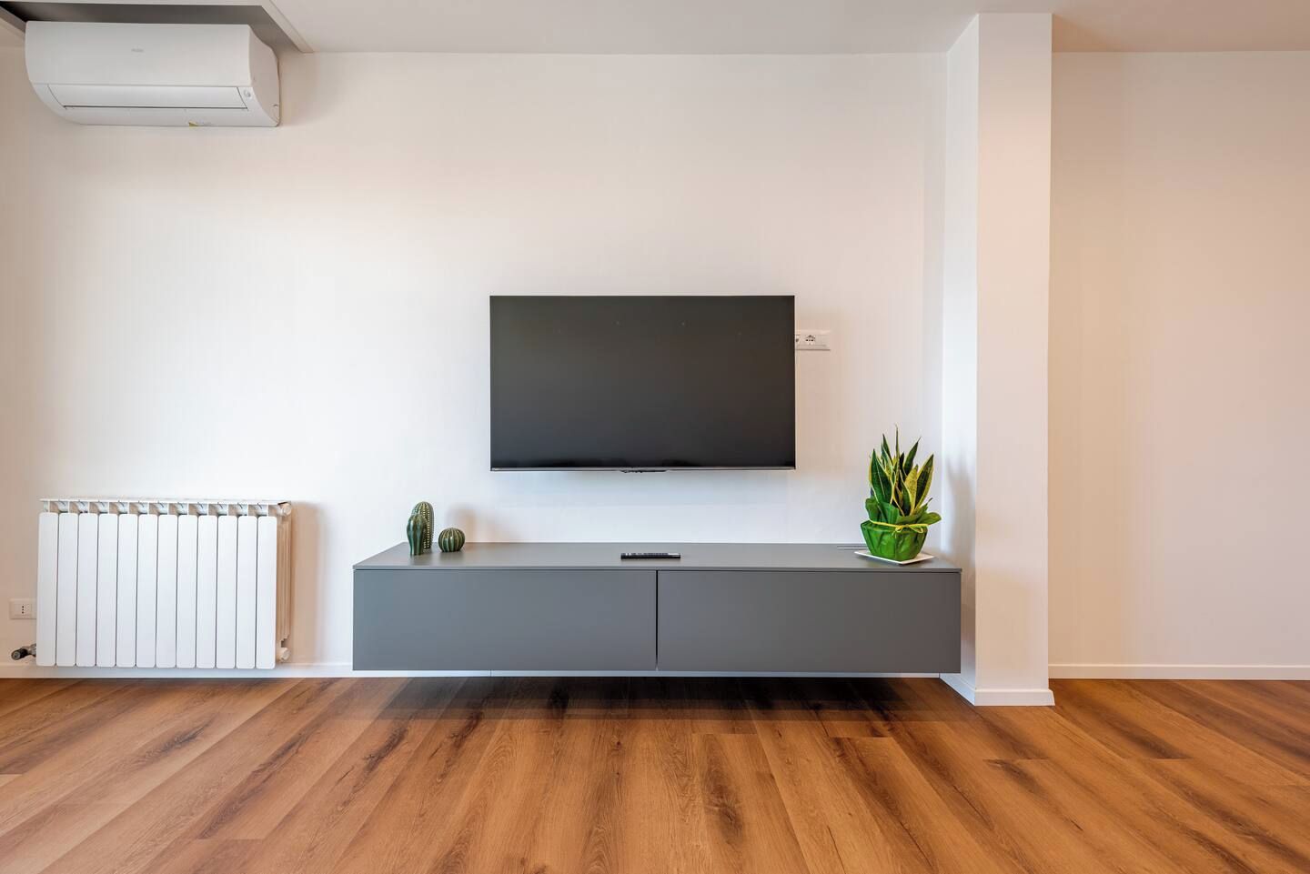 Un soggiorno moderno con una TV a schermo piatto montata su un mobile grigio, un termosifone e un pavimento in legno.