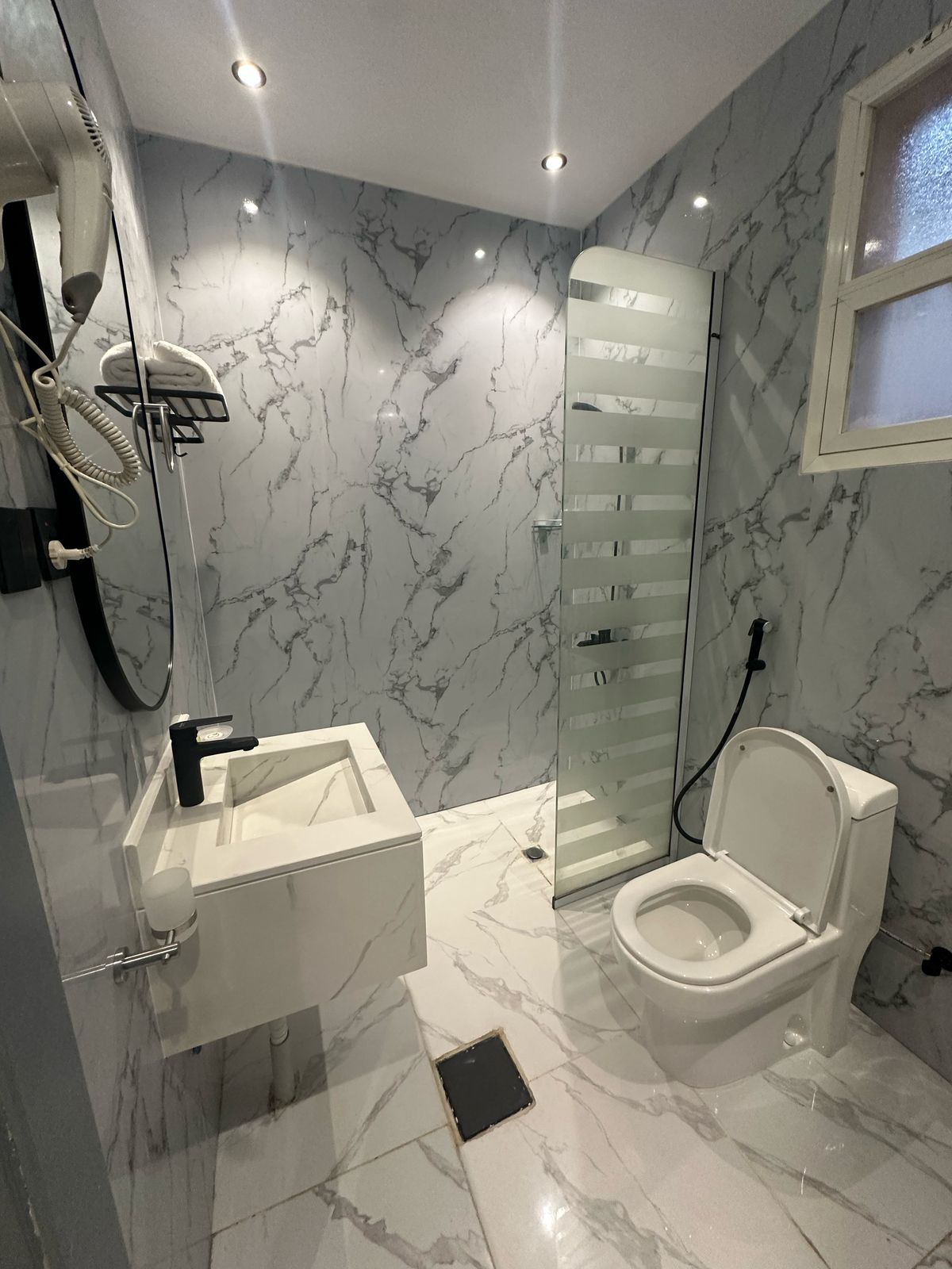 A modern bathroom with a marble floor, a white toilet, a glass shower enclosure, and a white sink with a black faucet.