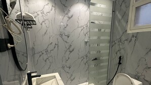 Un baño moderno con piso de mármol, inodoro blanco, cabina de ducha con mampara de vidrio y lavamanos blanco con grifo negro.