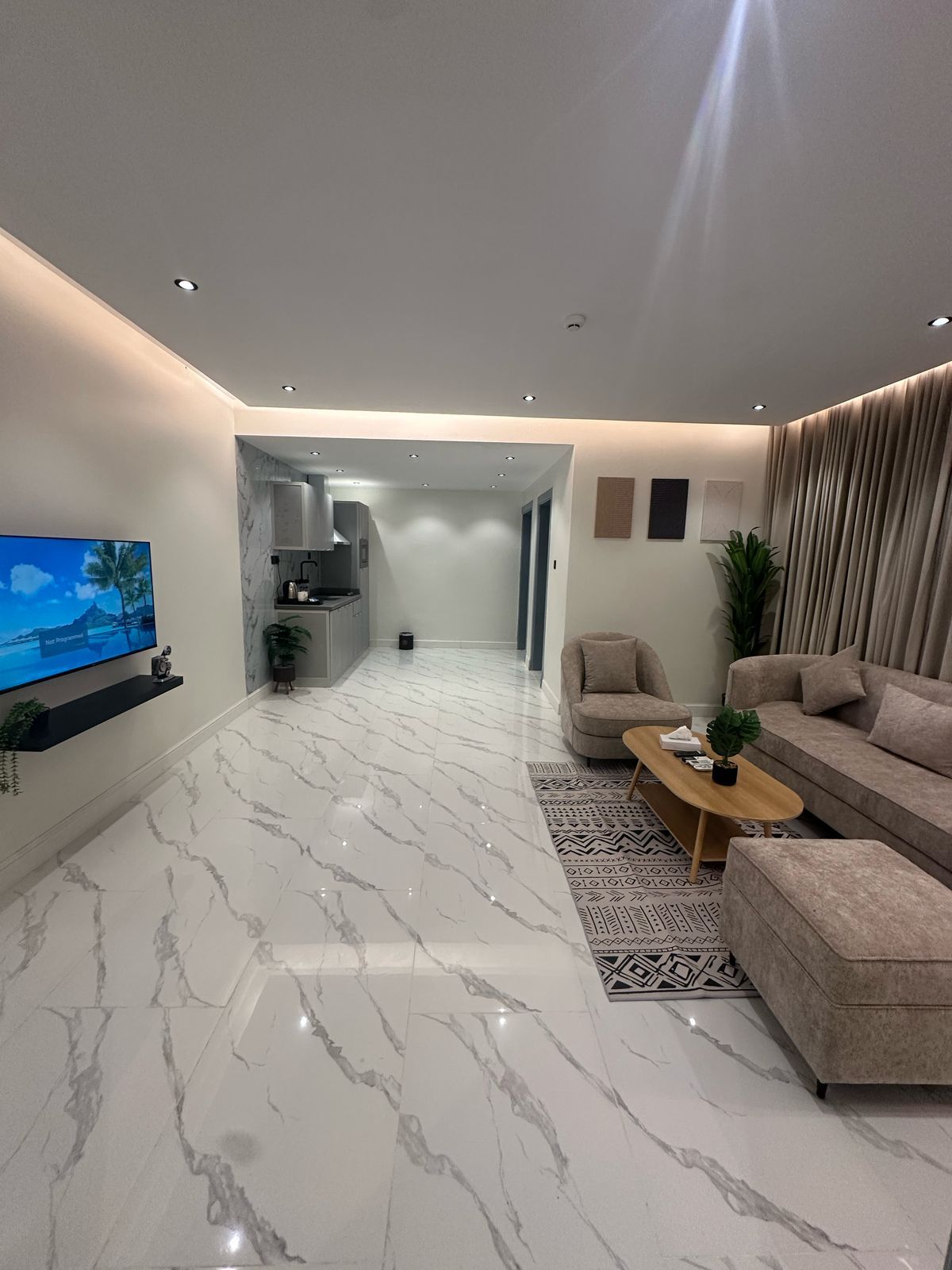 A modern living room with a flat-screen TV, a sofa, a coffee table, and a kitchen in the background.