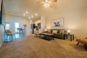 A spacious living area with a sofa, coffee table, and chairs, a kitchen in the background, and a ceiling fan. - The Winsome Western - HotTub - Fire Pit - PingPong (Lubbock)