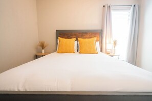 Una cama con ropa blanca y dos almohadas amarillas, un cabecero de madera, una mesita de noche con un jarrón de plantas secas y una ventana con cortinas.