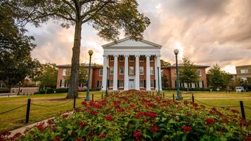 A building with a classical architectural style, featuring a central entrance with columns, surrounded by a well-maintained lawn with red flowers, and a cloudy sky in the background.