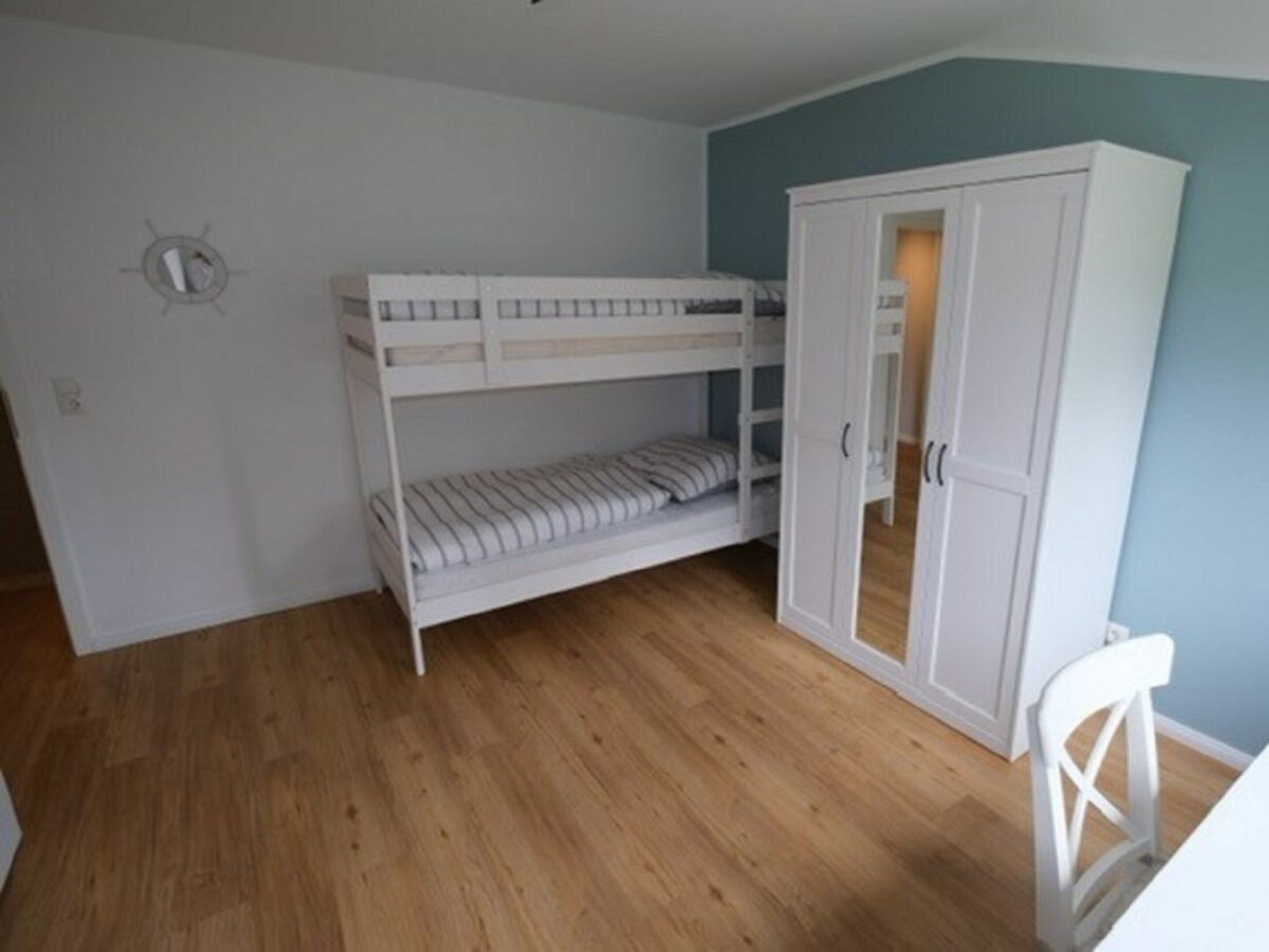 A bunk bed with a lower bunk, a white wardrobe, and a white chair in a room with wooden flooring and teal walls.