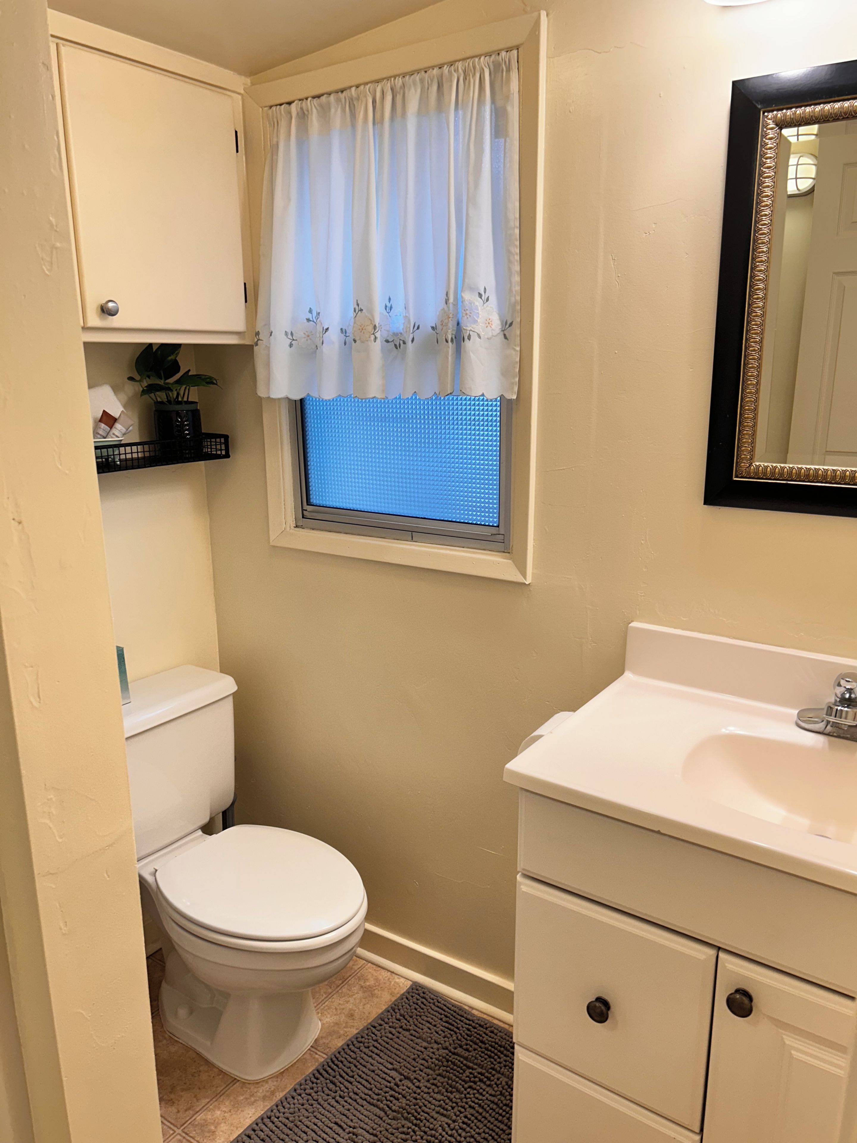 Un baño con inodoro, lavabo, espejo y ventana con cortina.
