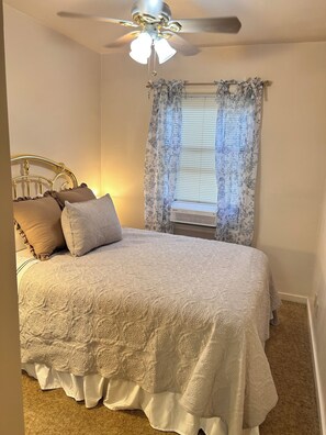 Un dormitorio con cama, ventilador de techo, ventana con cortinas florales y aire acondicionado de pared.