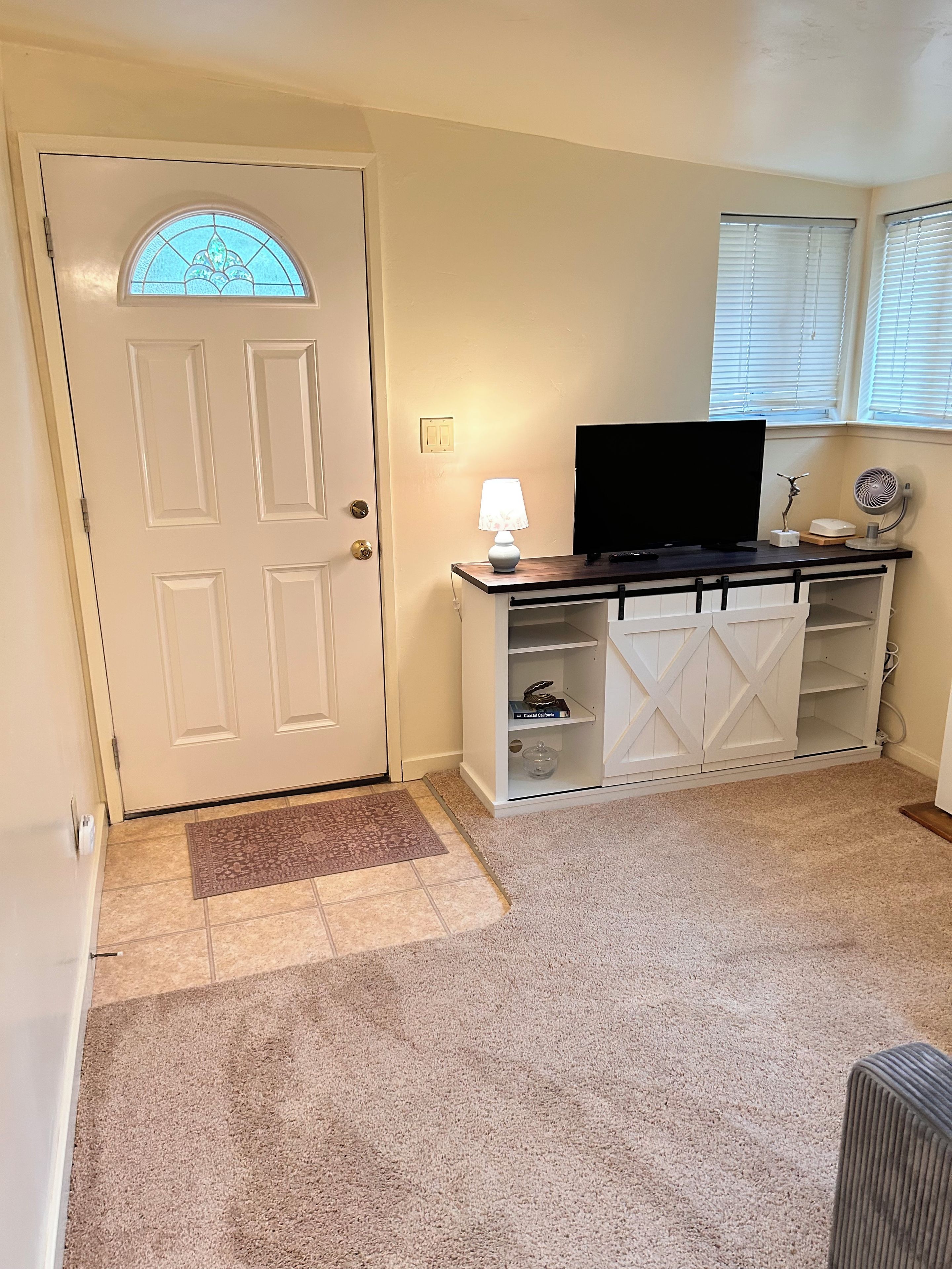 A room with a white door, a television on a wooden stand, a white cabinet with shelves, and a carpeted floor.